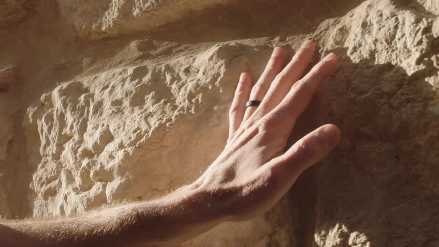 A man's hand gently touches and slides across a rough, ancient stone wall in the warm sunlight