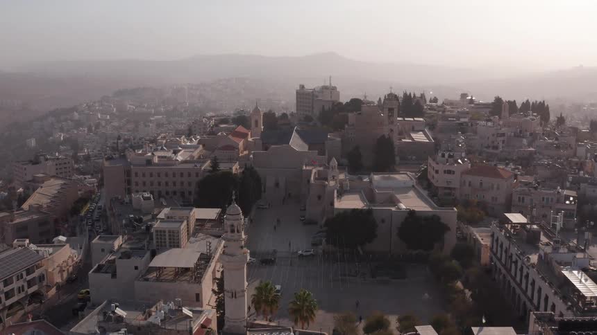 A descending aerial shot reveals the historic Church of the Nativity and Manger Square in the city of Bethlehem during a hazy sunrise
