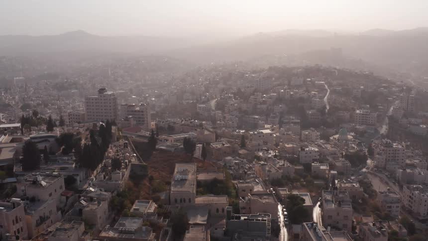 A slow and hazy aerial reveal of a sprawling Middle Eastern city nestled in rolling hills at sunrise