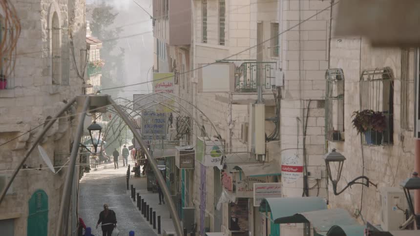 Pedestrians walk along a narrow, historic cobblestone street in an old Middle Eastern city as smoke drifts in the air