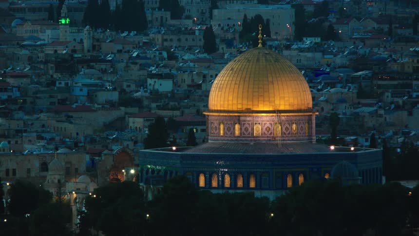 A rack focus shot reveals the illuminated golden Dome of the Rock against the cityscape of Jerusalem at dusk