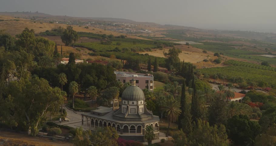 A serene, wide shot of the Church of the Beatitudes nestled among lush trees and overlooking the rolling hills of Galilee, Israel