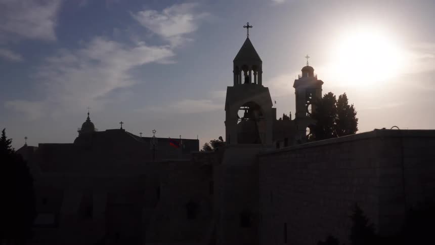 A slow aerial shot pulls back to reveal the historic Church of the Nativity in Bethlehem, silhouetted against the setting sun