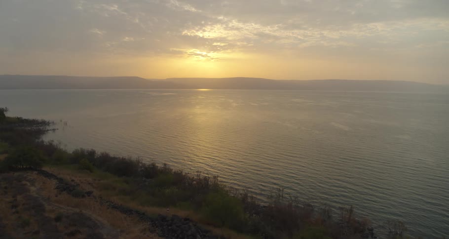 An aerial shot slowly glides over a rustic hillside to reveal the serene expanse of a vast lake during a golden sunset