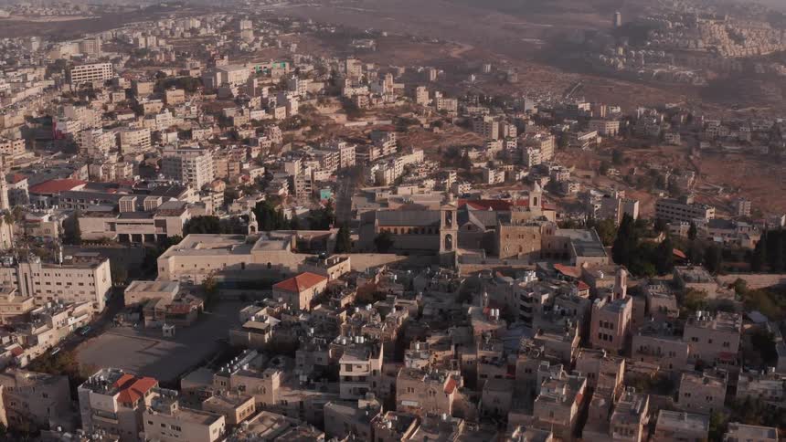 An aerial view slowly circles the historic Church of the Nativity, nestled within the sprawling cityscape of Bethlehem at sunrise