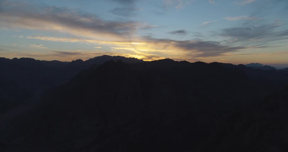A static wide shot captures the majestic silhouette of a mountain range against a colorful sky during a serene sunset