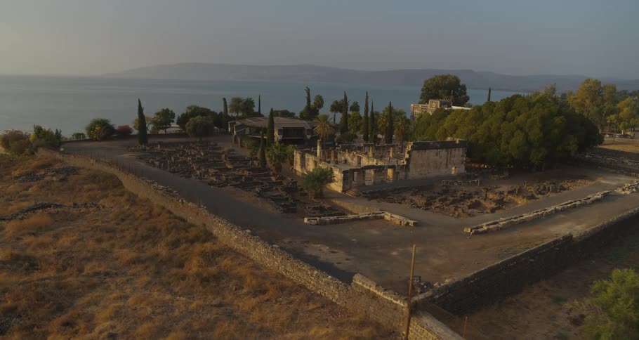 A slow aerial push-in reveals the ancient ruins of Capernaum on the shore of the Sea of Galilee at sunrise