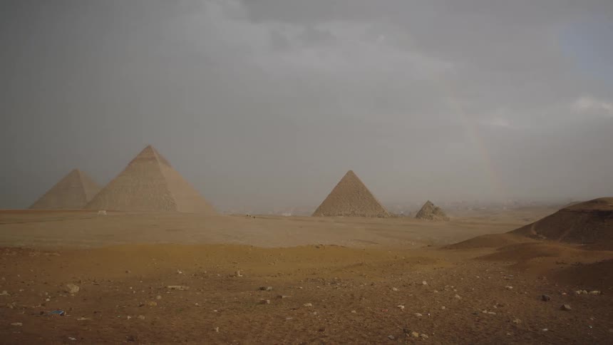A dramatic, wide shot of the Giza pyramids in the desert with a faint rainbow under a dark, stormy sky