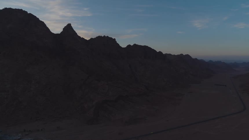 An epic aerial shot pans across a vast desert valley, revealing a silhouetted mountain range at dusk