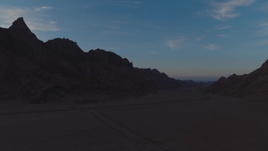 A slow aerial push-in shot over a vast desert valley with a lonely road flanked by mountain silhouettes at twilight