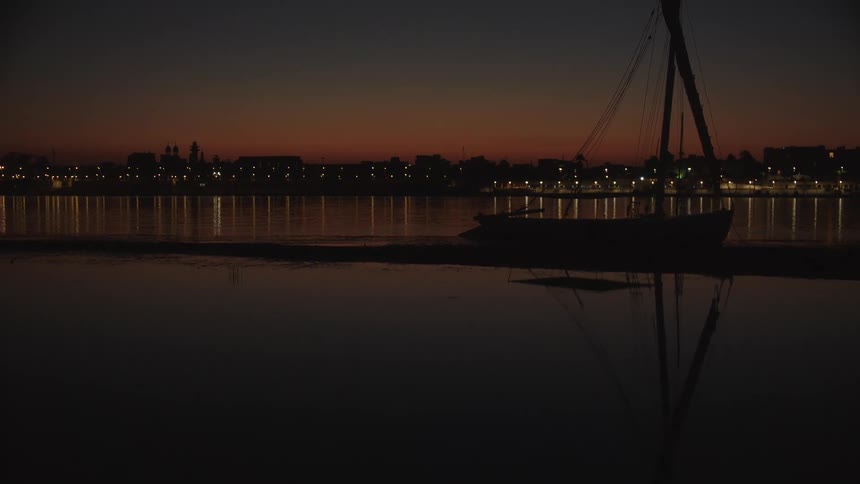 A silhouetted boat rests on the shore of a calm lake at dusk with a city skyline glowing in the background