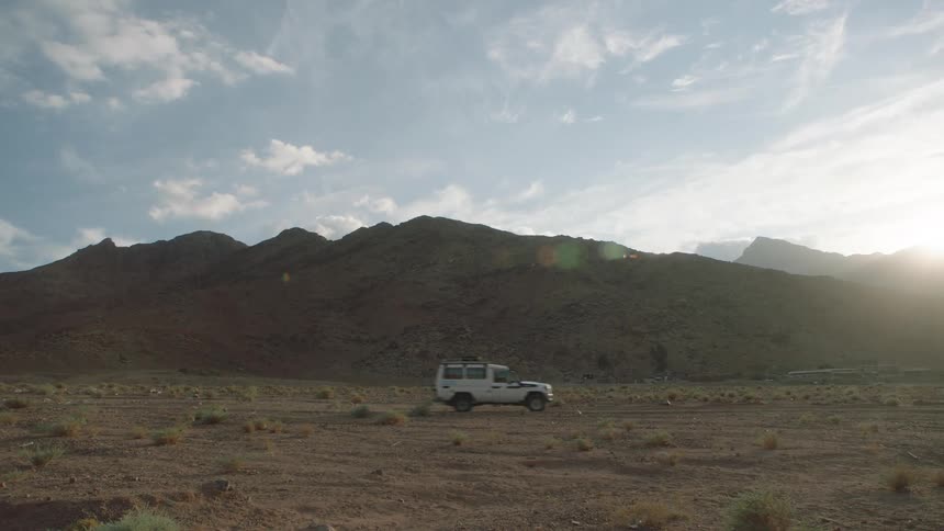 A white off-road vehicle drives across a dusty desert plain with a large mountain range in the background at sunset