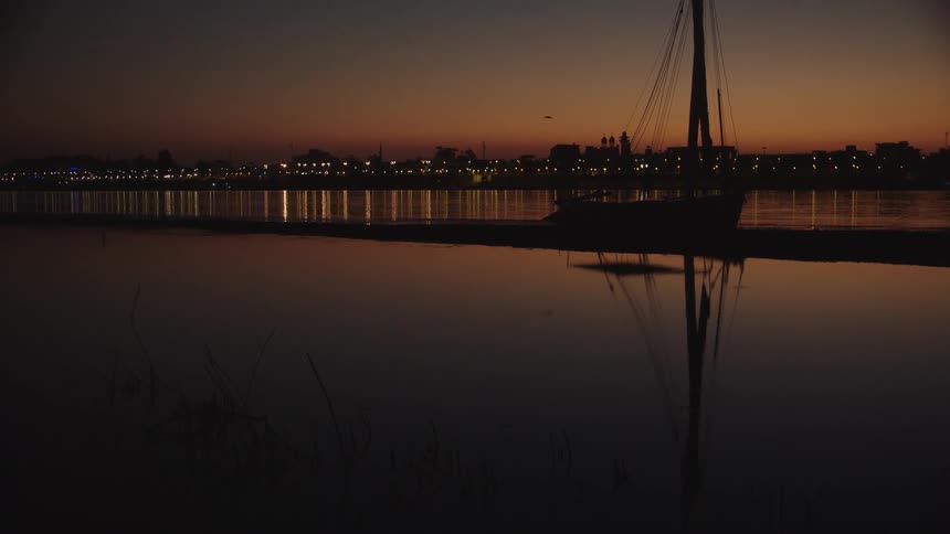 A tranquil scene of a silhouetted boat on a calm river at dusk, with its reflection and distant city lights shimmering on the water's surface