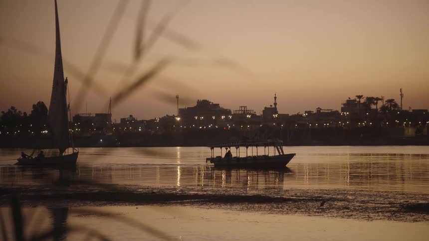Rack focus from riverside reeds reveals traditional boats on the Nile River at sunset, with the city lights of Luxor, Egypt twinkling in the distance