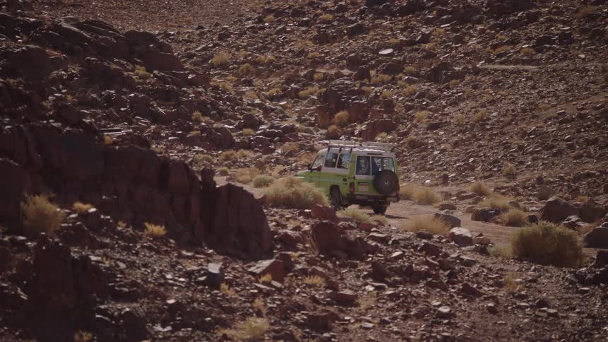Two 4x4 safari vehicles navigate a winding, rocky dirt road through a remote desert canyon