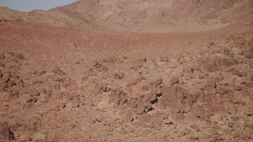 A lone white camper van sits at the base of a vast, rocky, red desert mountain range as the camera slowly reveals the epic scale of the landscape