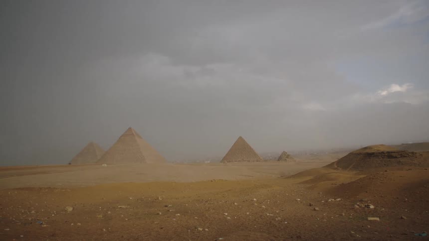 Ominous storm clouds gather over the Great Pyramids of Giza as rain begins to fall, revealing a faint rainbow in the sky