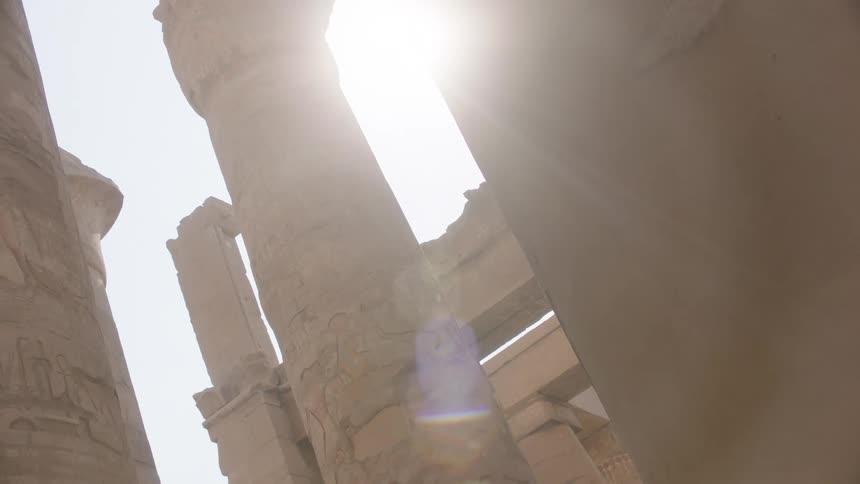 A low-angle view looking up at the colossal, hieroglyphic-covered columns of an ancient Egyptian temple as the sun flares brightly overhead