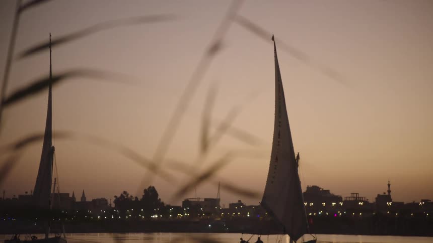 A calm evening on the Nile River in Egypt as traditional felucca sailboats drift past a city skyline at sunset, revealed in a beautiful rack focus shot