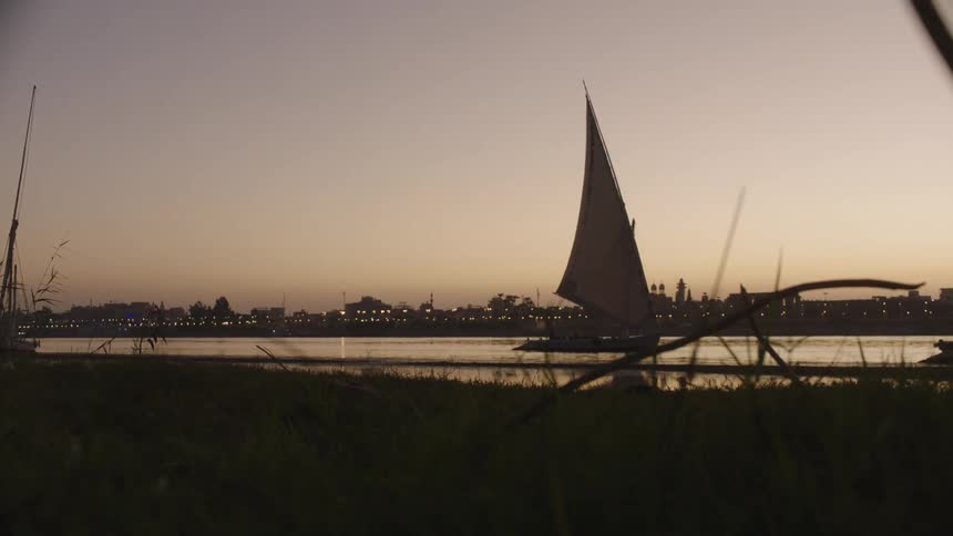 A traditional felucca sailboat peacefully glides across the river at dusk, with the glowing city skyline visible in the distance