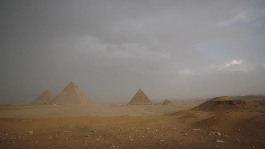 A faint rainbow forms in the hazy sky as dark storm clouds gather over the majestic Great Pyramids of Giza