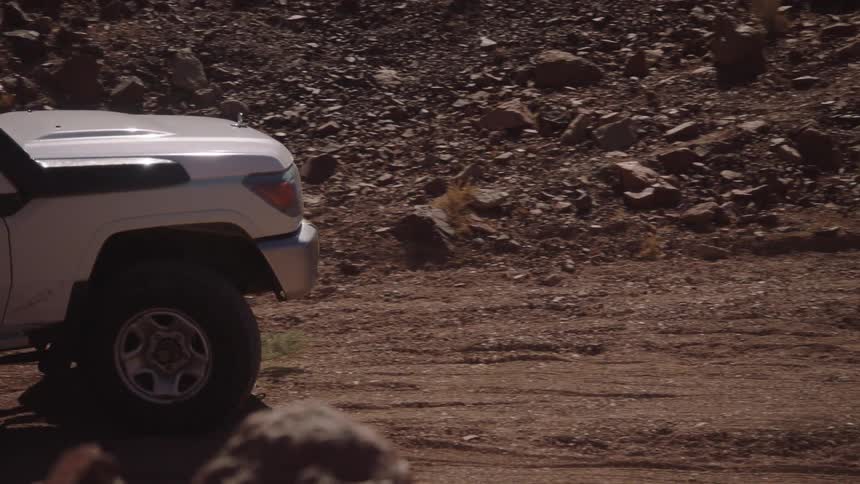 A white off-road truck drives slowly over a rugged dirt trail in a dry, rocky desert landscape