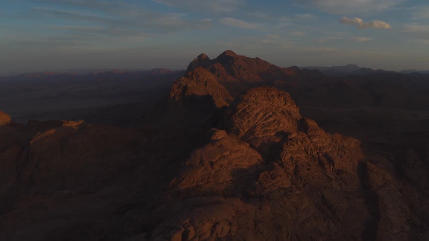 An epic aerial shot slowly pulls back to reveal a vast, rugged desert mountain range bathed in the warm light of sunrise