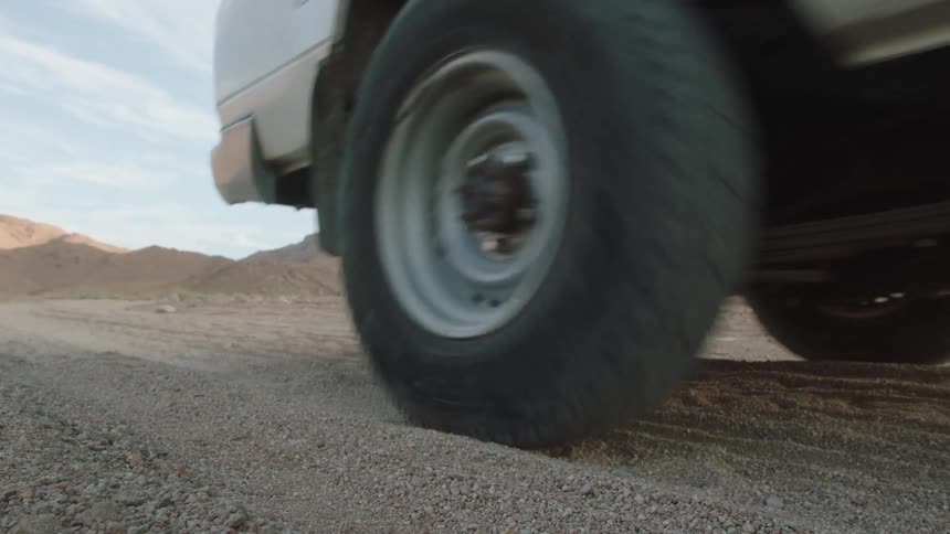 A slow-motion close-up of a car with a flat tire driving away on a dusty road in the desert