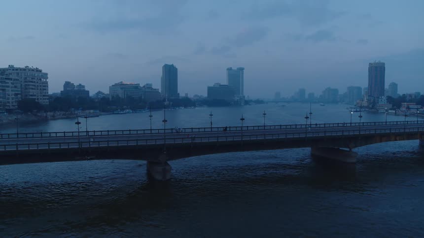 A wide, static shot of a bridge over a river at dusk, with the hazy city skyline in the background
