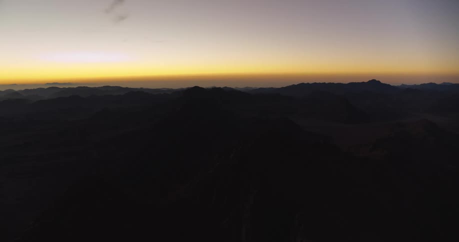 A static aerial shot captures the silhouette of a vast mountain range against the golden glow of a sunrise