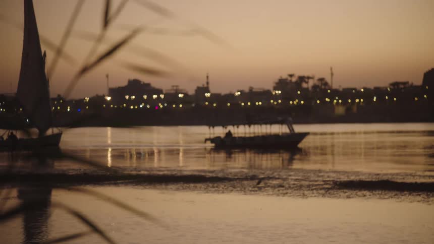 A beautiful focus pull from reeds in the foreground reveals boats on a tranquil river as city lights twinkle on the shore at dusk