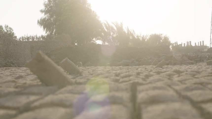 A low-angle, slow-motion rack focus shot reveals a desolate, hazy background beyond the sun-baked, cracked earth in the foreground
