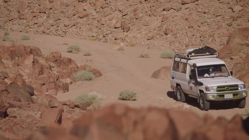 A white safari truck drives slowly along a dusty road through a rocky desert canyon