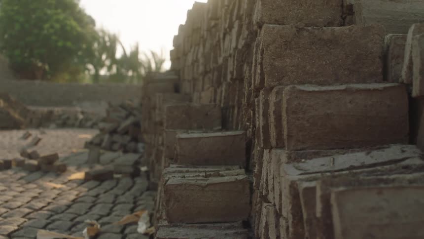 A slow pull-back shot reveals stacks of raw clay bricks drying in a brickyard as the bright sun creates a lens flare