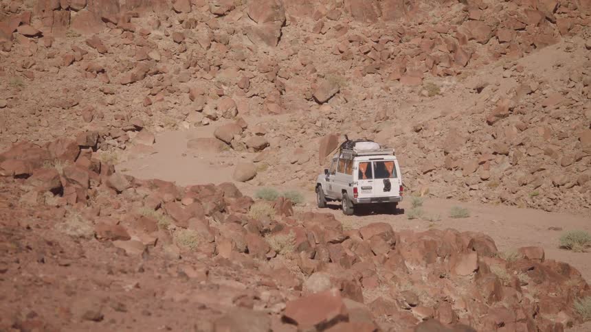 A white safari truck drives away on a dirt road through a remote, rocky desert canyon