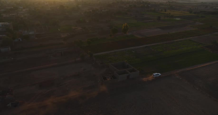 Aerial view of a white SUV kicking up dust as it travels along a dirt road through a rural village and farmland at sunset