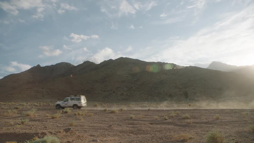 A white off-road vehicle kicks up dust as it drives through a rugged desert with mountains in the background