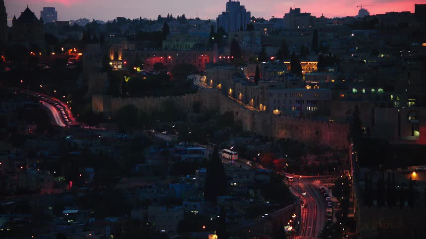 The Old City of Jerusalem glows with warm lights at dusk as traffic moves along the roads and a vibrant pink sunset colors the sky