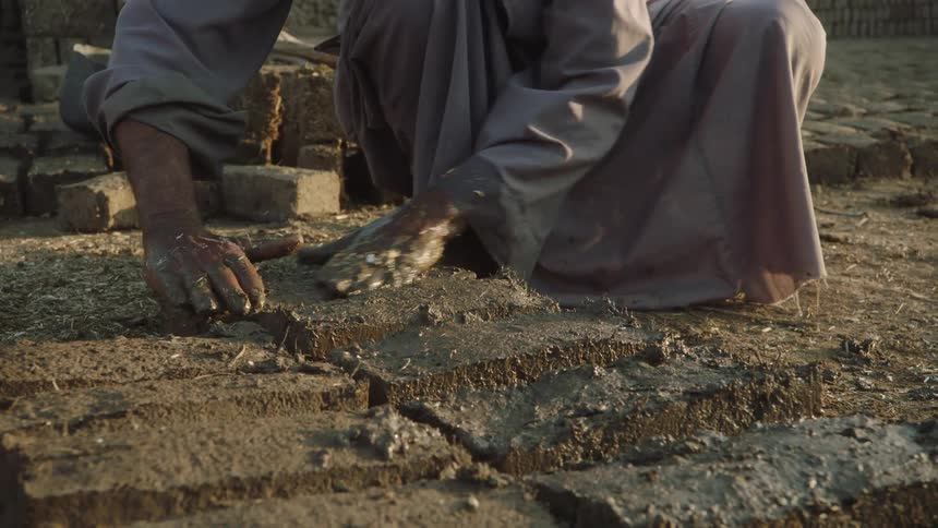 A man's hands covered in mud carefully shape handmade adobe bricks using a traditional wooden mold in slow motion