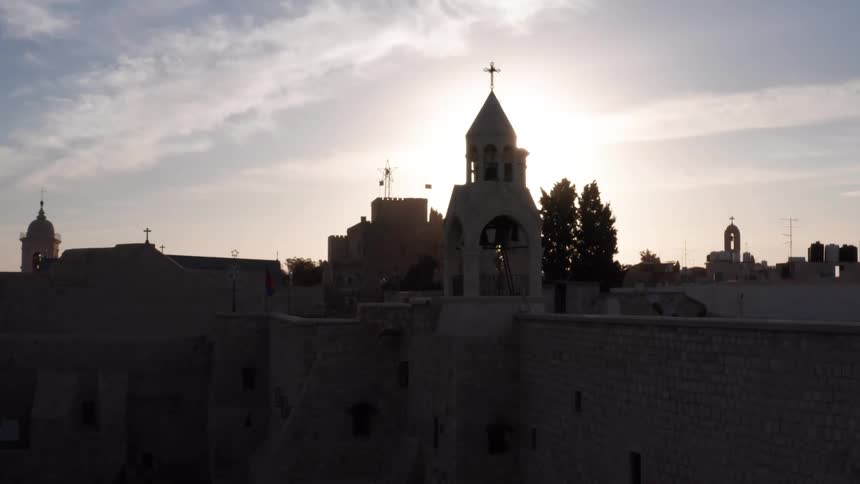A slow aerial reveal of a historic church bell tower silhouetted against the bright morning sun in the ancient city of Bethlehem