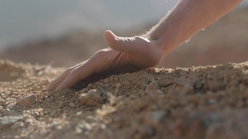 A slow-motion close-up shows a hand gently touching down and gripping a handful of dry, rocky soil