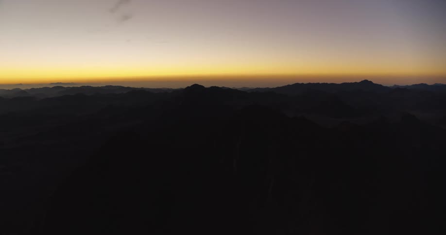 A tranquil, wide shot shows the first light of dawn glowing on the horizon behind a majestic, silhouetted mountain range