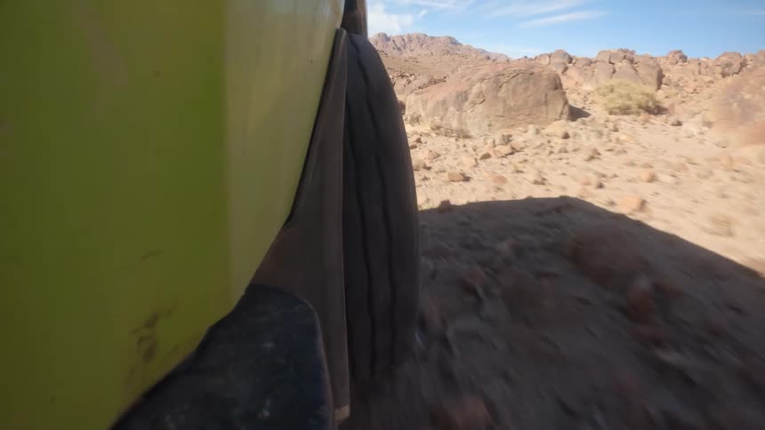 A point-of-view shot from the side of a lime green off-road vehicle shows its tire navigating a challenging rocky desert trail