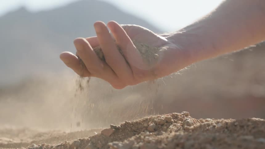 In a sun-drenched arid landscape, a hand slowly releases a handful of dry earth, letting it sift through the fingers in a mesmerizing slow-motion display