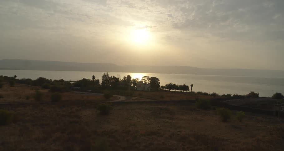 A slow aerial push-in reveals a historic church on the tranquil shores of the Sea of Galilee as the sun rises over the water