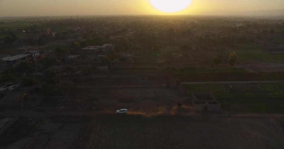 An aerial view captures a white vehicle kicking up dust as it drives through a rural village landscape at sunset