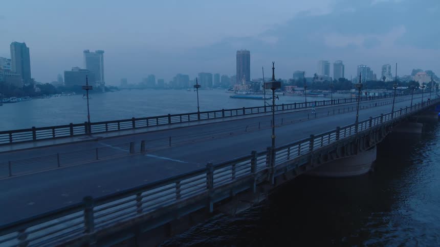 Aerial drone footage follows a man walking and a man jogging across a deserted bridge over a river with a city skyline in the background at dawn