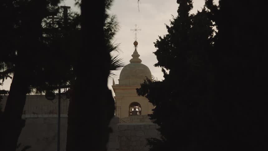 A stone church steeple with a cross stands against a dusky sky, viewed through silhouetted trees