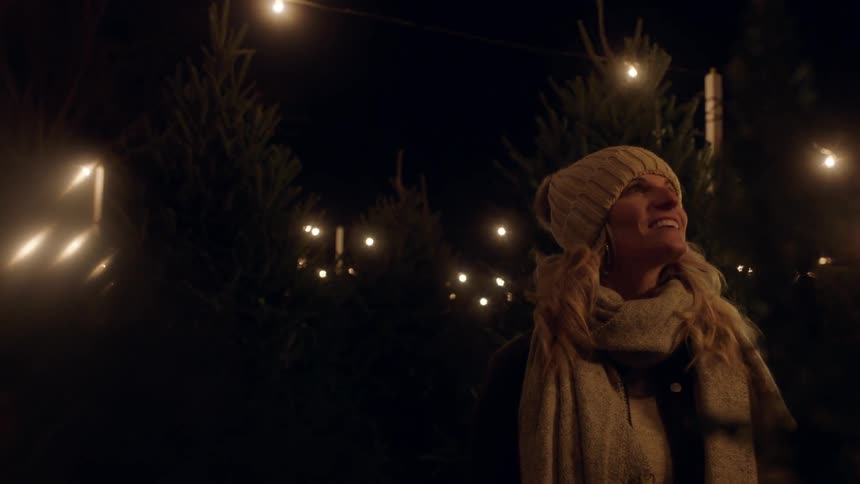 A woman smiles outdoors at night while wearing a scarf and hat