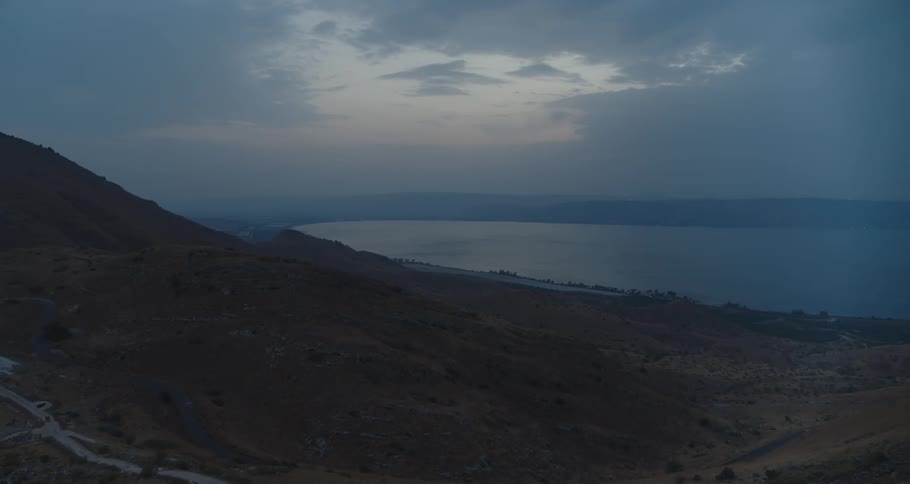 A slow and majestic aerial drone shot flies over the arid hills towards a vast lake under a moody, cloudy sky at twilight